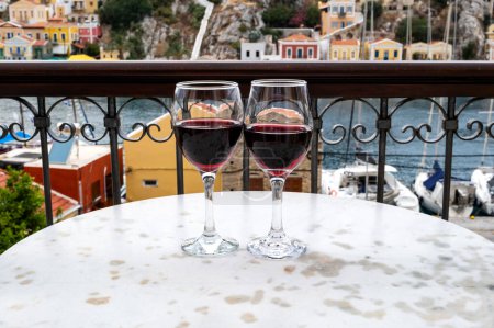 Foto de Dos copas de vinos tintos en un bistró de mármol y una vista del puerto principal de la isla de Symi, Grecia - Imagen libre de derechos