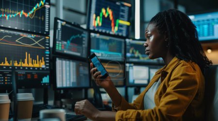 In this compelling image, a black woman confidently navigates the complexities of the stock market, surrounded by a high-tech office environment. With her smartphone in hand, she seamlessly integrates technology into her trading strategy, while multi