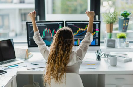 In this dynamic image, a woman celebrates a victorious moment in her trading journey, her arms raised in triumph at her desk. Behind her, screens display stock market data and charts, a testament to her astute decision-making and strategic prowess. W