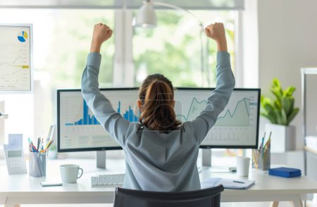 In this triumphant image, a woman sits at her desk, arms raised in celebration after achieving a significant win on the stock market through strategic trading. Her face radiates with joy and satisfaction as she basks in the glow of her success. Behin