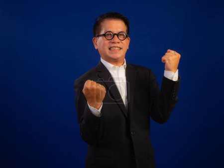 Téléchargez les photos : Portrait de heureux d'âge moyen asiatique confiant homme d'affaires réussi portant costume célébrant sur fond bleu en studio, en regardant la caméra - en image libre de droit