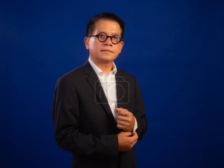 Téléchargez les photos : Portrait de intelligent d'âge moyen asiatique confiant homme d'affaires réussi portant costume debout sur fond bleu en studio, à la recherche de leadership professionnel attrayant et positif - en image libre de droit