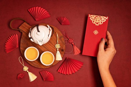 Téléchargez les photos : Décoration du Nouvel An chinois. Traditionnel Nouvel An lunaire plat posé avec du thé vert, ventilateurs en papier rouge, main femme tenant poche rouge. Vue du dessus - en image libre de droit
