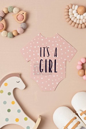 Photo for Baby shower, gender reveal party. It's a girl message over paper cut onesie. Flatlay, top view on a beige pastel background. Newborn gifts. Invitation, celebration, greeting card idea - Royalty Free Image