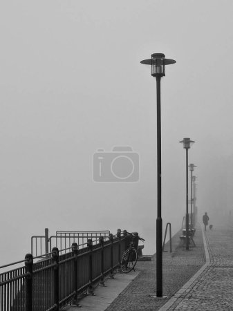 Foggy morning, woman walking with dog on the quay of Danube River in Passau, Bavaria, Germany. The mysterious cobblestone street in the city, day with fog in black and white.