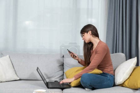 Young woman sitting on sofa reading promotional code on smartphone typing on laptop computer for discount price on the web site of the internet store.