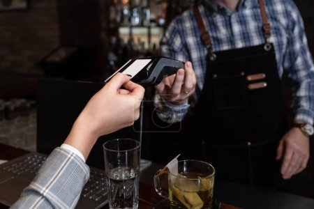 Female business person hand holding credit card paying for drink in cafeteria with contact less wi-fi bank card to the waiter who hold paying machine. Modern payment without touching concept.
