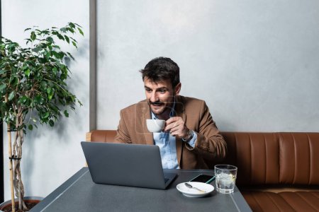 Photo for Young successful self employed freelancer man working from cafeteria on laptop computer while drinking coffee and enjoying his freedom. New small business owner businessperson working from coffee shop - Royalty Free Image