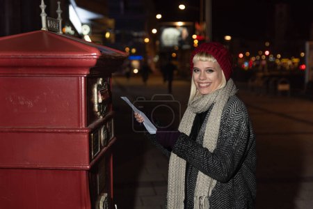 Téléchargez les photos : Night portrait of young beautiful hipster girl with cap and scarf in coat near mailbox putting letter envelope sending love message to her boyfriend - en image libre de droit