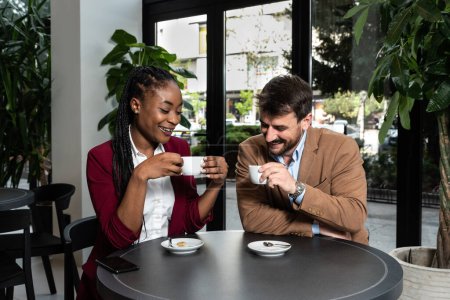 Foto de Young man and African American woman sitting in cafeteria drinking coffee flirting talking laughing and seducing outside working hours and colleague in restaurant away from busy street - Imagen libre de derechos