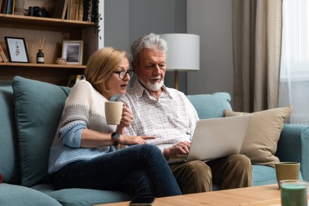 Photo for Happy middle aged family couple relaxing on sofa, using computer web surfing information shopping together at home. Elderly man showing laptop apps to retired wife, older generation with tech concept. - Royalty Free Image
