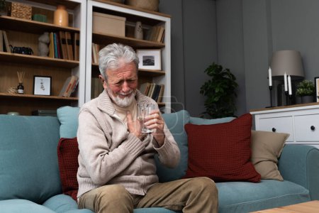 Photo for Senior older man getting chest pain in living room, heart attack holding his hand on the chest sitting on sofa at home. Health care older people issue - Royalty Free Image
