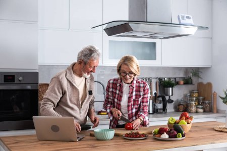 Téléchargez les photos : Heureux vieux couple d'âge moyen utilisant un ordinateur portable pour préparer une salade de légumes diète saine à la maison ensemble, rechercher des recettes, commander des achats en ligne, regarder des cours de cuisine dans la cuisine. - en image libre de droit