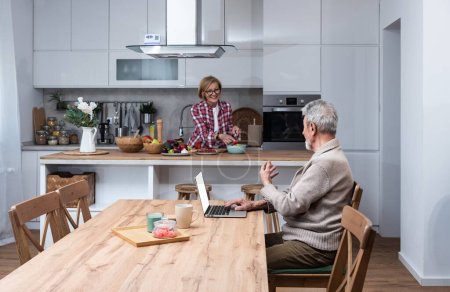 Glücklicher älterer älterer Mann Ehemann Fernarbeit oder Fernstudium online aus dem Home Office mit Laptop-Computer, während ihre ältere Frau Frühstück in der Küche kocht. Familienpaar mittleren Alters zu Hause.