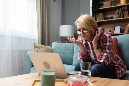 Worried middle-aged 70s lady sit on sofa in living room read negative unexpected news on laptop, anxious mature woman rest on couch feel distressed with unpleasant bad message on computer