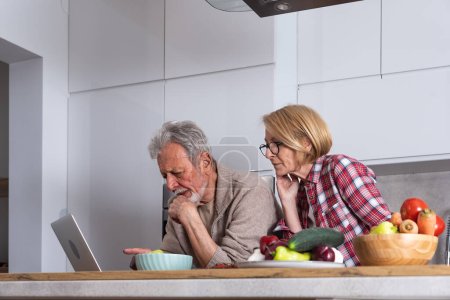 Téléchargez les photos : Couple aîné cuisiner des aliments sains ensemble à partir de légumes biologiques à la maison dans la cuisine tout en lisant et en préparant la recette d'Internet sur ordinateur portable - en image libre de droit