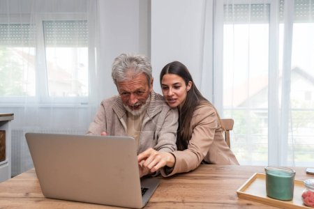 Photo for Daughter and elderly father or grandfather sitting at the table at home daughter teaches him to use laptop computer and surf the internet to find information easily entertainment and read news - Royalty Free Image