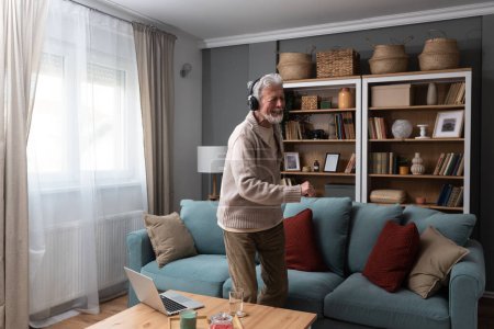 Hombre feliz mayor bailando en la sala de estar en casa con música en el portátil y auriculares inalámbricos, celebrando la jubilación y terminando el trabajo. Anciano pensionista danza masculina