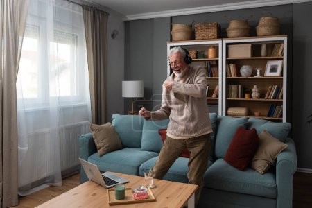 Glücklicher älterer Mann, der zu Hause im Wohnzimmer mit Musik auf Laptop und drahtlosen Kopfhörern tanzt, den Ruhestand feiert und die Arbeit beendet. Seniorentanz