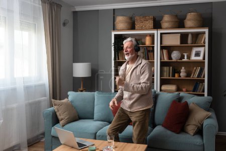 Téléchargez les photos : Homme heureux âgé dansant dans le salon à la maison avec de la musique sur ordinateur portable et écouteurs sans fil, célébrer la retraite et le travail de finition. Pensionné âgé danse masculine - en image libre de droit