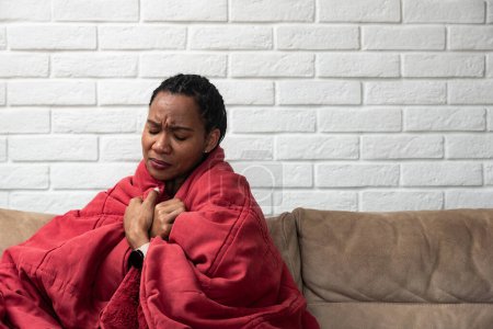 Téléchargez les photos : Malade jeune femme africaine se sentant froid recouvert d'une couverture assis sur le canapé, malade fille noire frissonnant froid réchauffement à la maison enveloppé avec plaid, aucun problème de chauffage central, concept de grippe fièvre température - en image libre de droit