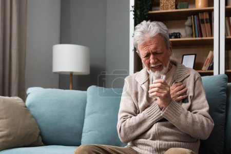 Photo for Senior older man getting chest pain in living room, heart attack holding his hand on the chest sitting on sofa at home. Health care older people issue trying to drink water from glass - Royalty Free Image