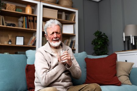Photo for Senior older man getting chest pain in living room, heart attack holding his hand on the chest sitting on sofa at home. Health care older people issue trying to drink water from glass - Royalty Free Image