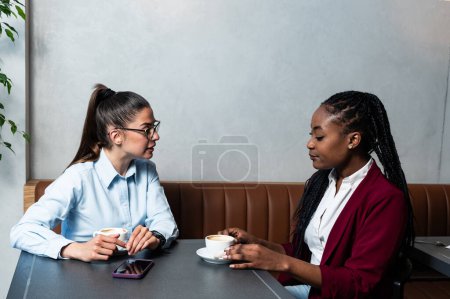 Foto de Dos jóvenes colegas empresarias tomando un descanso en la cafetería cercana tomando café hablando de la vida privada, para conocerse mejor. Miembros del personal empleados de la empresa sentados en la cafetería - Imagen libre de derechos
