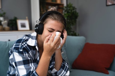 Photo for Young sad girl sitting alone at home on sofa listening to sad music on wireless headphones crying and wiping her nose with a paper tissue. Memories and missing concept. - Royalty Free Image