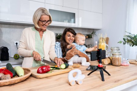 Foto de Familia de las Tres Generaciones. La abuela, la madre y el bebé graban un vlog de cocina o podcast mientras cortan verduras para una comida vegetariana saludable en la cocina y lo transmiten en línea. Preparación de alimentos veganos - Imagen libre de derechos