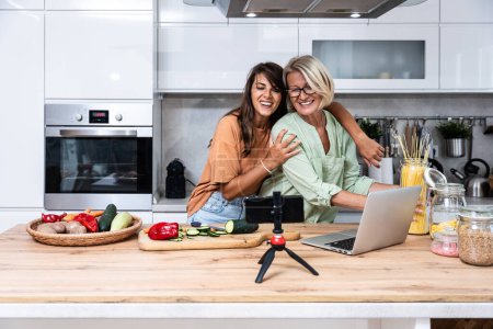 Foto de La abuela y su hija graban un vlog de cocina o podcast mientras cortan verduras para una comida vegetariana saludable en la cocina y lo transmiten en línea. Preparación de alimentos veganos - Imagen libre de derechos