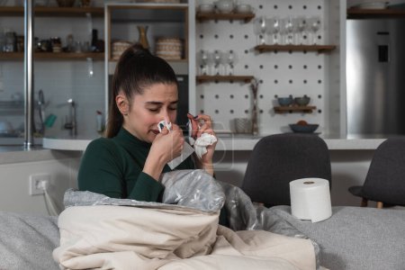 Photo for Young sick woman wrapped in a blanket wipes her nose with paper tissues, got sick because she didn't have money to pay for heating in her home or breakdown in the city heating network system. - Royalty Free Image