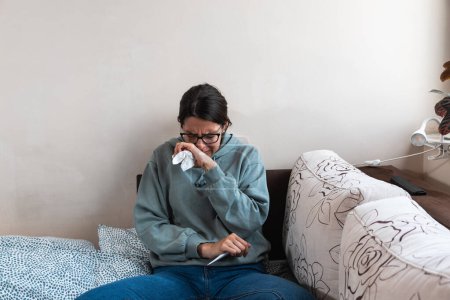 Photo for Young sick woman wrapped in a blanket wipes her nose with paper tissues, got sick because she didn't have money to pay for heating in her home or breakdown in the city heating network system. - Royalty Free Image