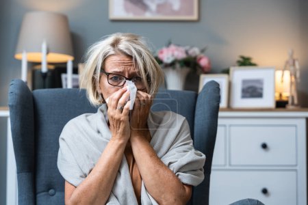 Photo for Older senior woman wrapped in a blanket wipes her nose with paper tissues, got sick because she didn't have money to pay for heating in her home or breakdown in the city heating network system. - Royalty Free Image