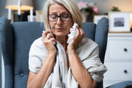 Photo for Older senior woman wrapped in a blanket wipes her nose with paper tissues, got sick because she didn't have money to pay for heating in her home or breakdown in the city heating network system. - Royalty Free Image