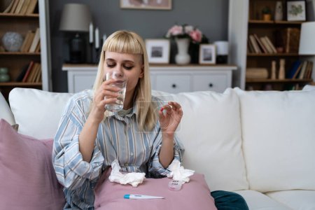 Young sick woman drinking medical pills and water after high body temperature and fever. Sick female feeling better with new medicine vitamins, sitting at home and taking her therapy.