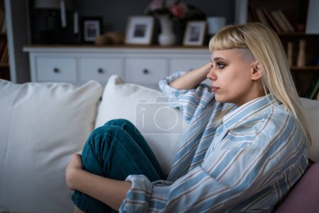 Caucasian sad woman sitting at the sofa with depression. Young student female suffering from mental burnout, feeling depressed, relationship difficulties, sits alone in silence at her home.