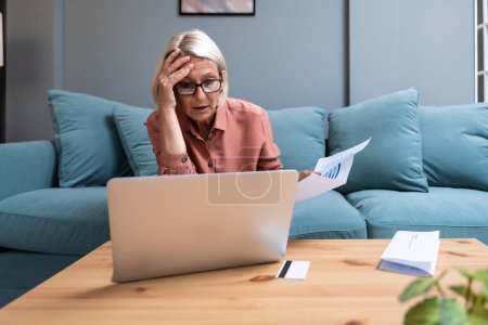 Shocked concerned senior accountant woman finding financial failure, mistake, bankruptcy, sitting at laptop, calculator, paper bills, covering face in panic attack, staring at screen