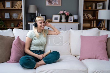 Young woman relaxing on a sofa at home. Listening music on wireless headphones and reading book. Book lover female enjoy free time and weekend activities for relaxation at her cozy apartment