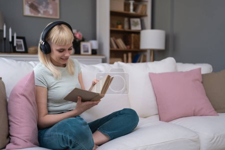 Young woman relaxing on a sofa at home. Listening music on wireless headphones and reading book. Book lover female enjoy free time and weekend activities for relaxation at her cozy apartment