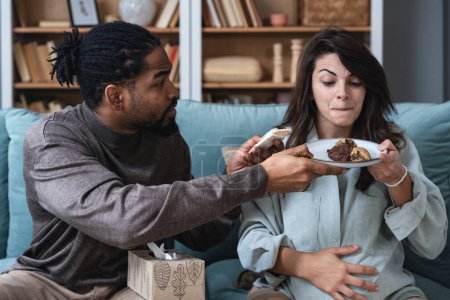Pregnancy support and fun. Young couple man and pregnant woman sitting at home teasing with sweet food forbidden by doctor. Male and female support and love during pregnancy period enjoy together.