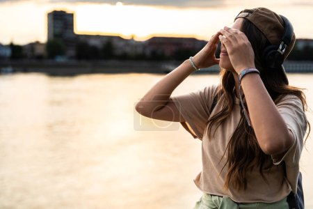 Young hipster millennial woman sitting on riverside looking at city listening music on wireless headphones enjoying moment, life and sunset. Happy independent girl enjoy freedom and life concept