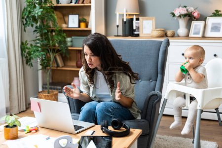 Young mother, business woman having video call on laptop from home while taking care of her baby sitting in tall baby chair. Female business owner on maternity leave must work online from home.