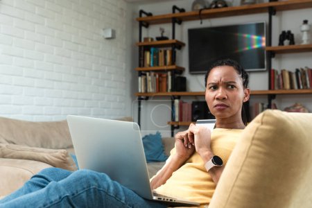 Confused woman sitting on couch holds credit card use laptop looking at device screen having debt problems, transaction failed, money withdraw impossible, insecure online payment or scam fraud concept