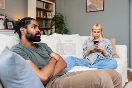 Téléchargez les photos : Young couple sitting on sofa at home, woman ignoring man, typing text messages on smartphone, male looking in distance with deep thoughts. Relationship difficulties. Two people problems - en image libre de droit