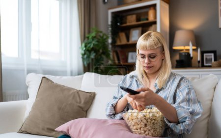 Young happy freshly divorced or recently single woman after toxic relationship breakup sitting alone at home on sofa, eating popcorn and watching movie or television in her new apartment she rented.
