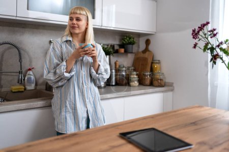 Young happy freshly divorced or recently single woman after toxic relationship breakup standing at kitchen in new apartment she rent drinking coffee or tea enjoying new beginnings and morning silence
