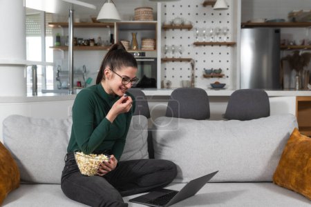 Young happy freshly divorced or recently single woman after toxic relationship breakup sitting alone at home on sofa, eating popcorn and watching movie on laptop in her new apartment she rented.