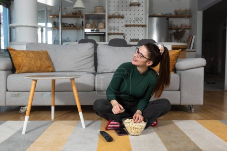 Young happy freshly divorced or recently single woman after toxic relationship breakup sitting alone at home on floor, eating popcorn and watching movie or television in her new apartment she rented.