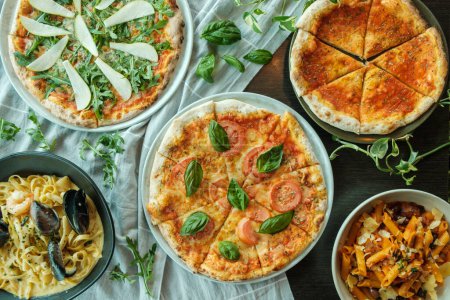 Foto de Mesa de madera llena de plato italiano tradicional, pizzas y pasta con hojas verdes alrededor de la mesa, vista superior, cena y menú de almuerzo - Imagen libre de derechos